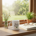 White mug with 'Mommy Gardener' text on a windowsill with plants and a book.