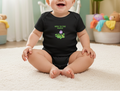 Baby wearing a black onesie with a design and text, sitting on a carpeted floor.
