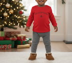 Child wearing a red 'Merry Christmas' sweater with a tree design, standing in front of a decorated Christmas tree and presents.