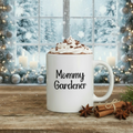 White mug with 'Mommy Gardener' text, hot chocolate with whipped cream, and cinnamon sticks on a wooden surface with a festive background.