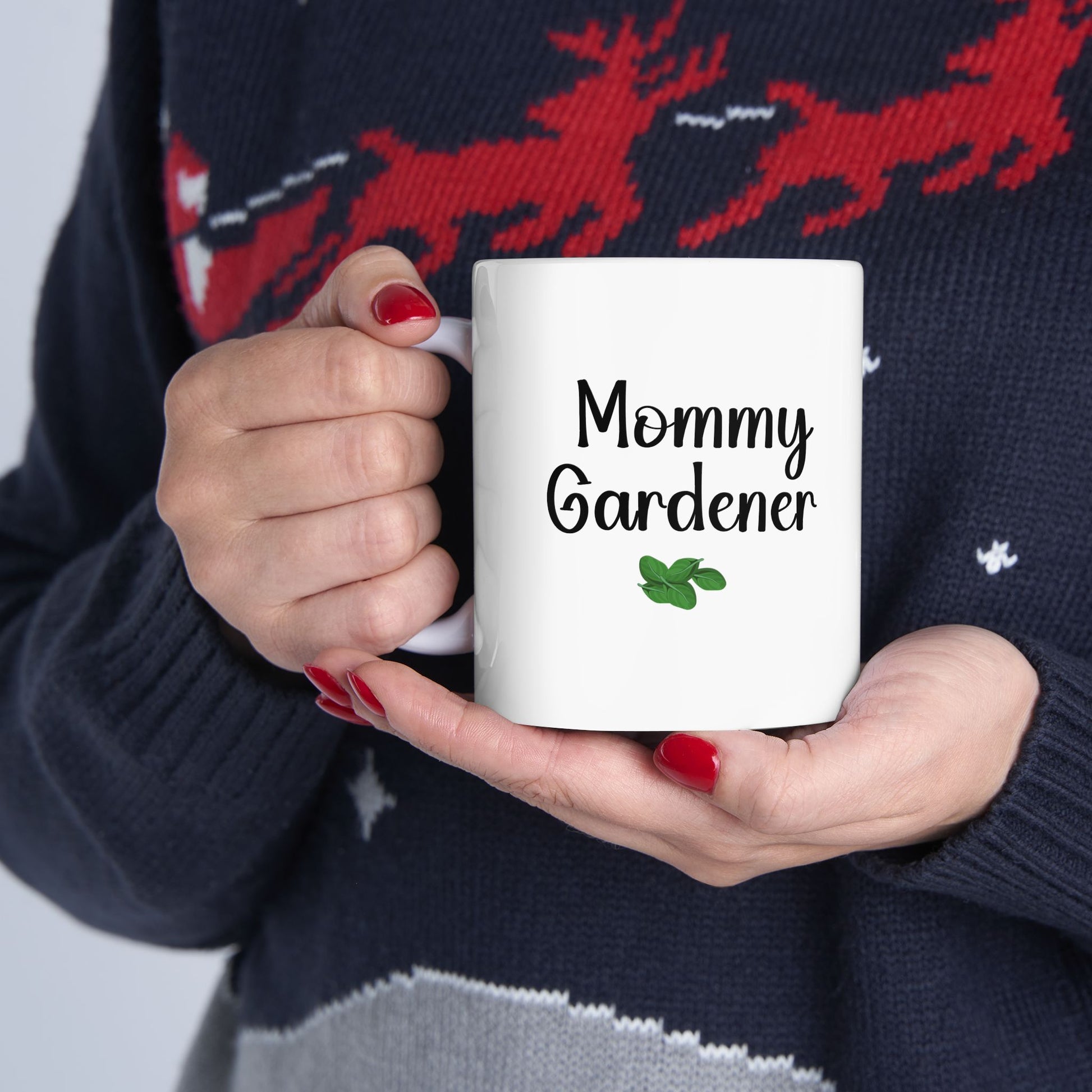 Person holding a white mug with 'Mommy Gardener' text, wearing a dark sweater with red deer design.