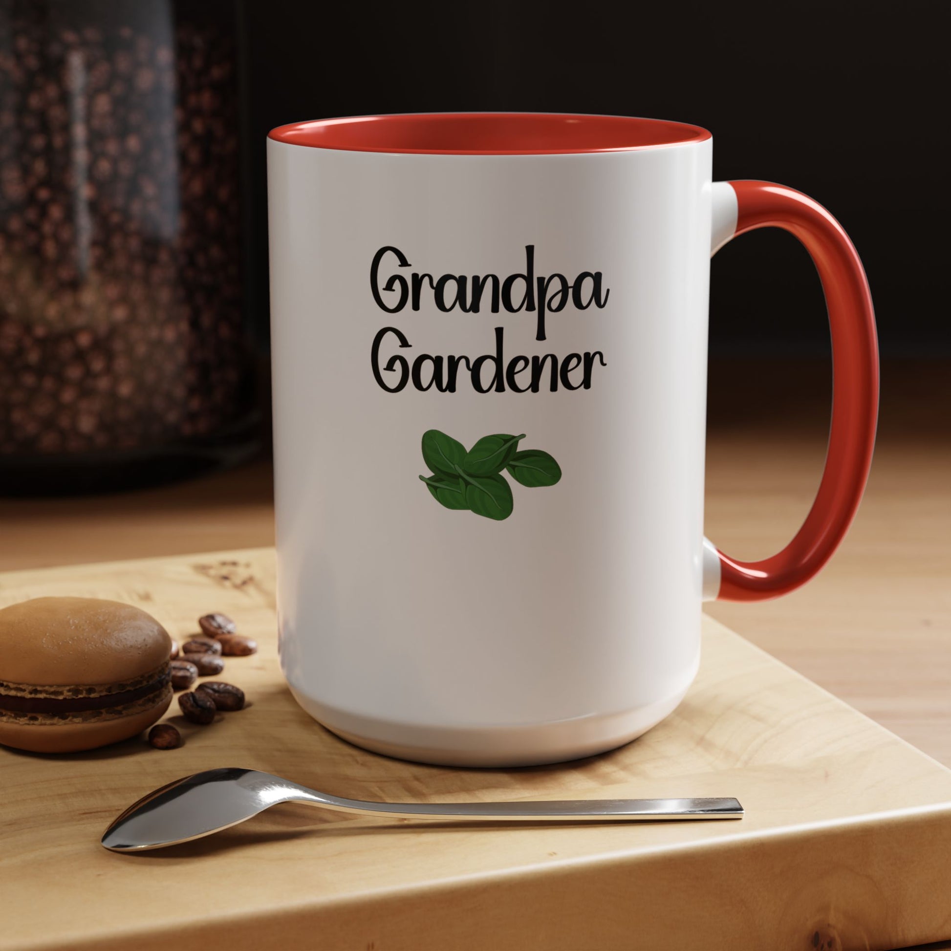 White mug with red interior and handle, featuring 'Grandpa Gardener' text and a leaf design, on a wooden surface with coffee beans and a macaron.