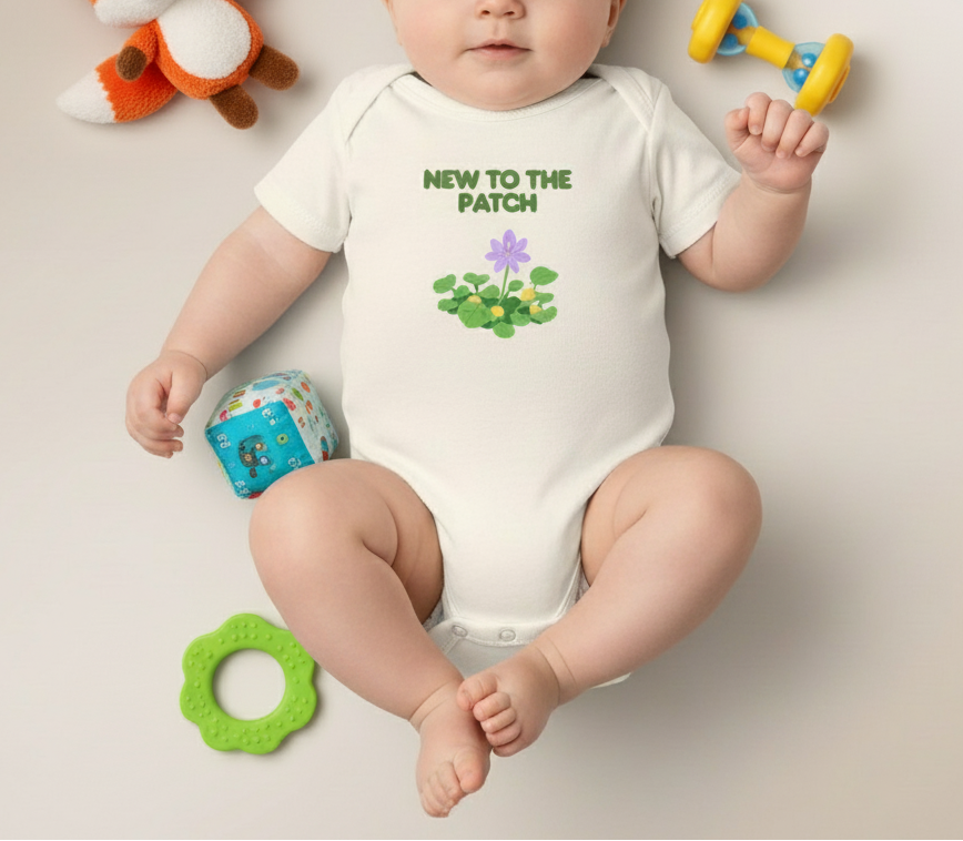 Baby wearing a onesie with 'New to the Patch' text and flowers, surrounded by toys on a plain background