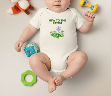 Baby wearing a onesie with 'New to the Patch' text and flowers, surrounded by toys on a plain background