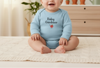 Baby wearing a light blue onesie with 'Baby Gardener' text and a tomato graphic, sitting on a beige blanket.