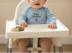 Baby in a high chair wearing a blue 'Baby Gardener' shirt with a pink flower graphic.