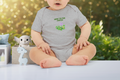 Baby wearing a gray onesie with a green logo and text, sitting on a surface with a blurred outdoor background.