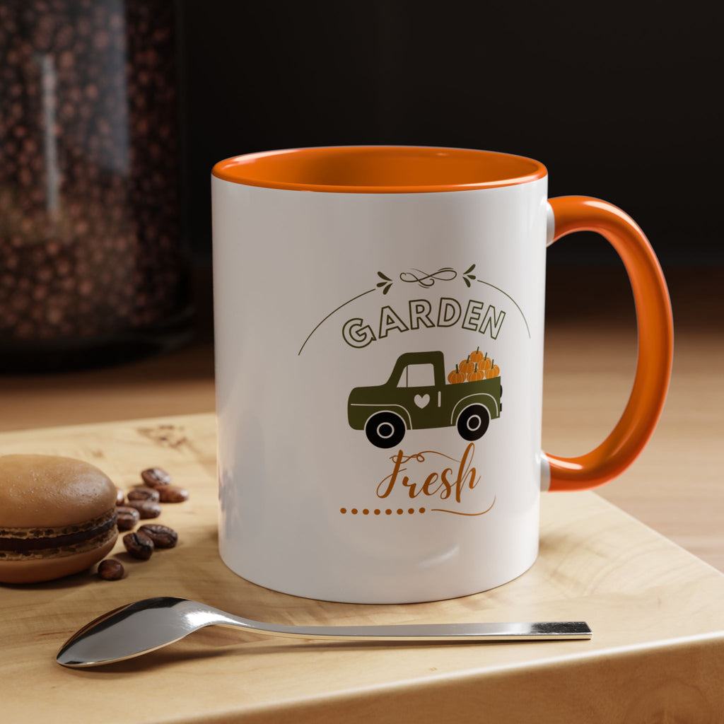 White mug with orange interior and handle, featuring a truck and 'Garden Fresh' text, on a wooden surface with coffee beans and a spoon.