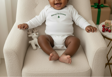 Baby sitting in a chair wearing a 'Merry Christmas' onesie with a reindeer toy next to them.