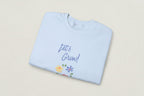 Folded light blue sweatshirt with 'Let's Grow!' text and floral designs on a white background