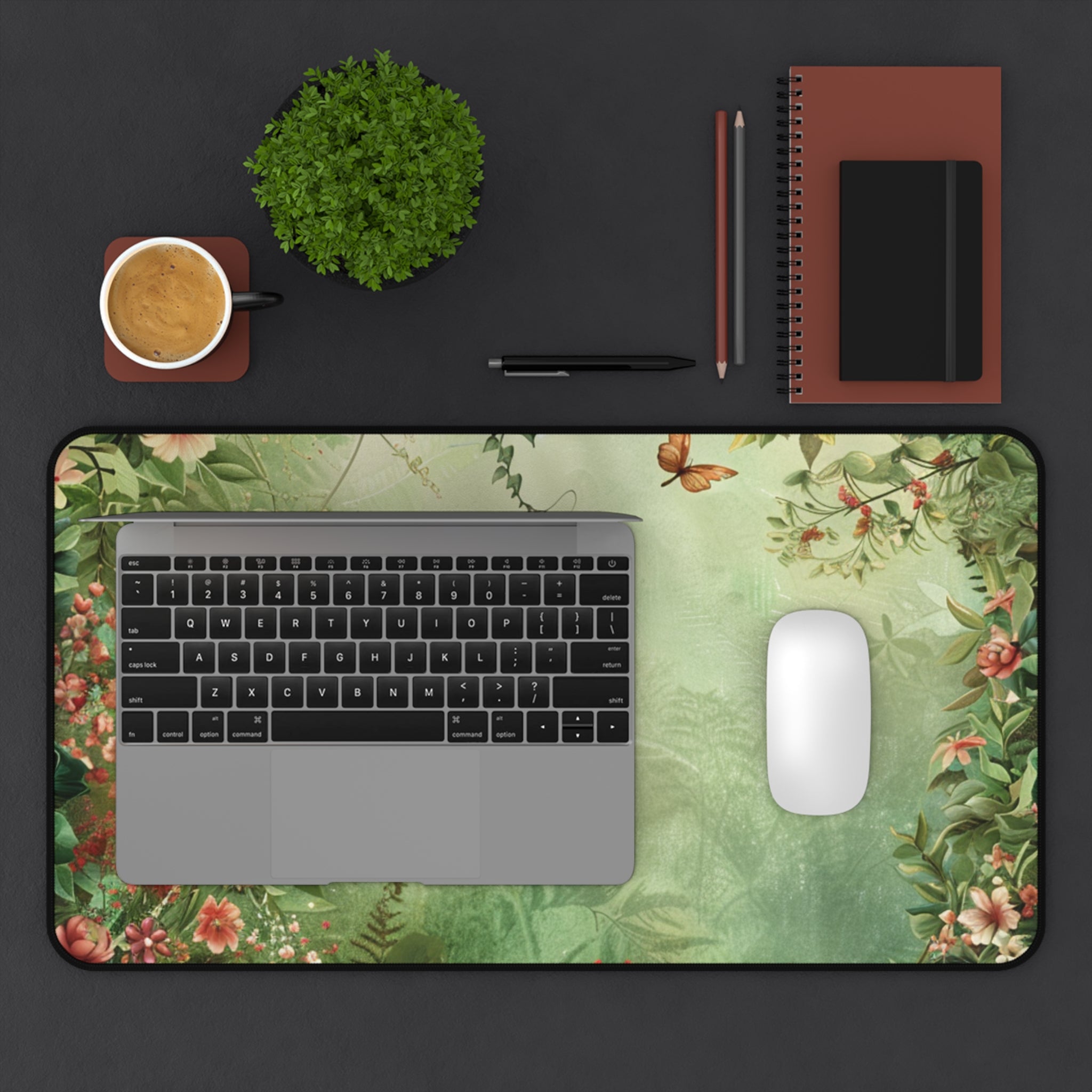 Laptop on a floral desk mat with a cup, plant, pen, and notebook on a dark surface
