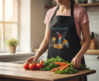 Person wearing a black apron with a gardening design, standing in a kitchen with vegetables on a wooden table.