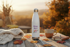 White water bottle with 'Grow, Girl!' text on a wooden table with a mug and leaves during sunset.