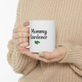 Person holding a white mug with 'Mommy Gardener' text and a leaf graphic, wearing a beige sweater.