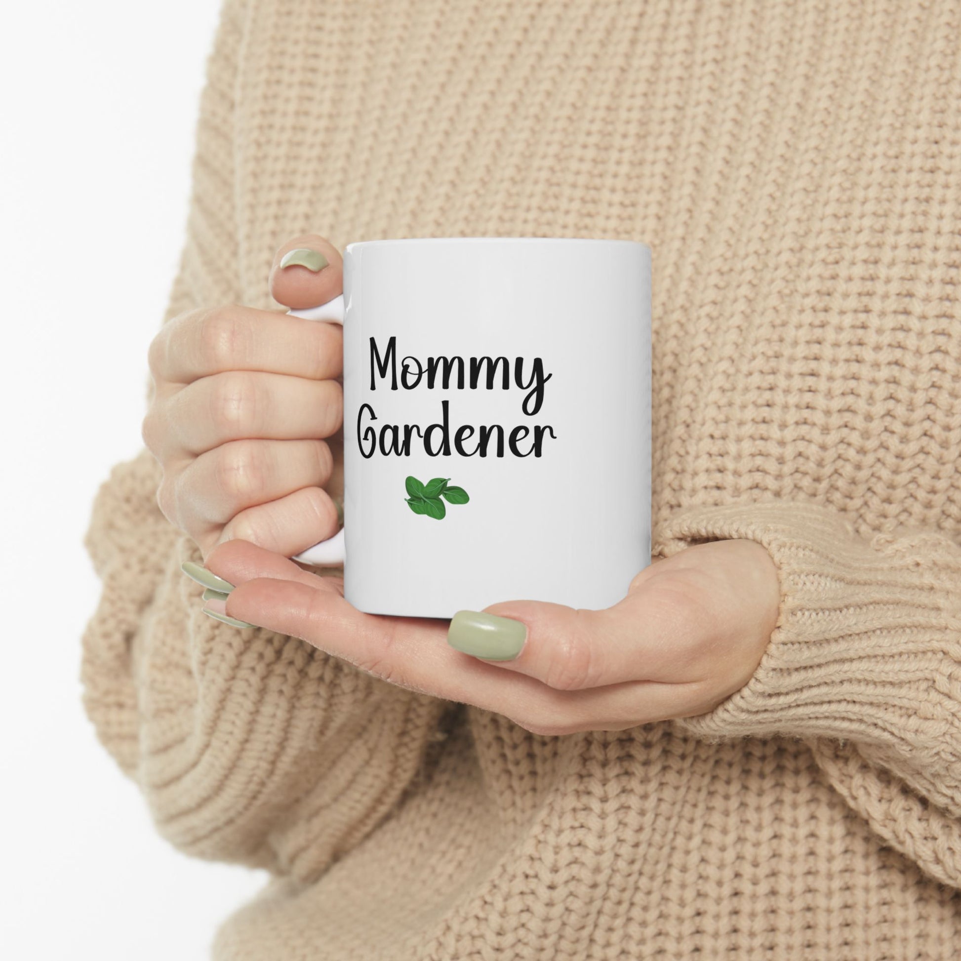 Person holding a white mug with 'Mommy Gardener' text and a leaf graphic, wearing a beige sweater.
