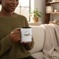 Person holding a mug with a design of broccoli, tomato, and pepper in a cozy living room.