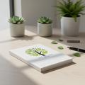 White notebook with green tree design on a light surface with potted plants in the background