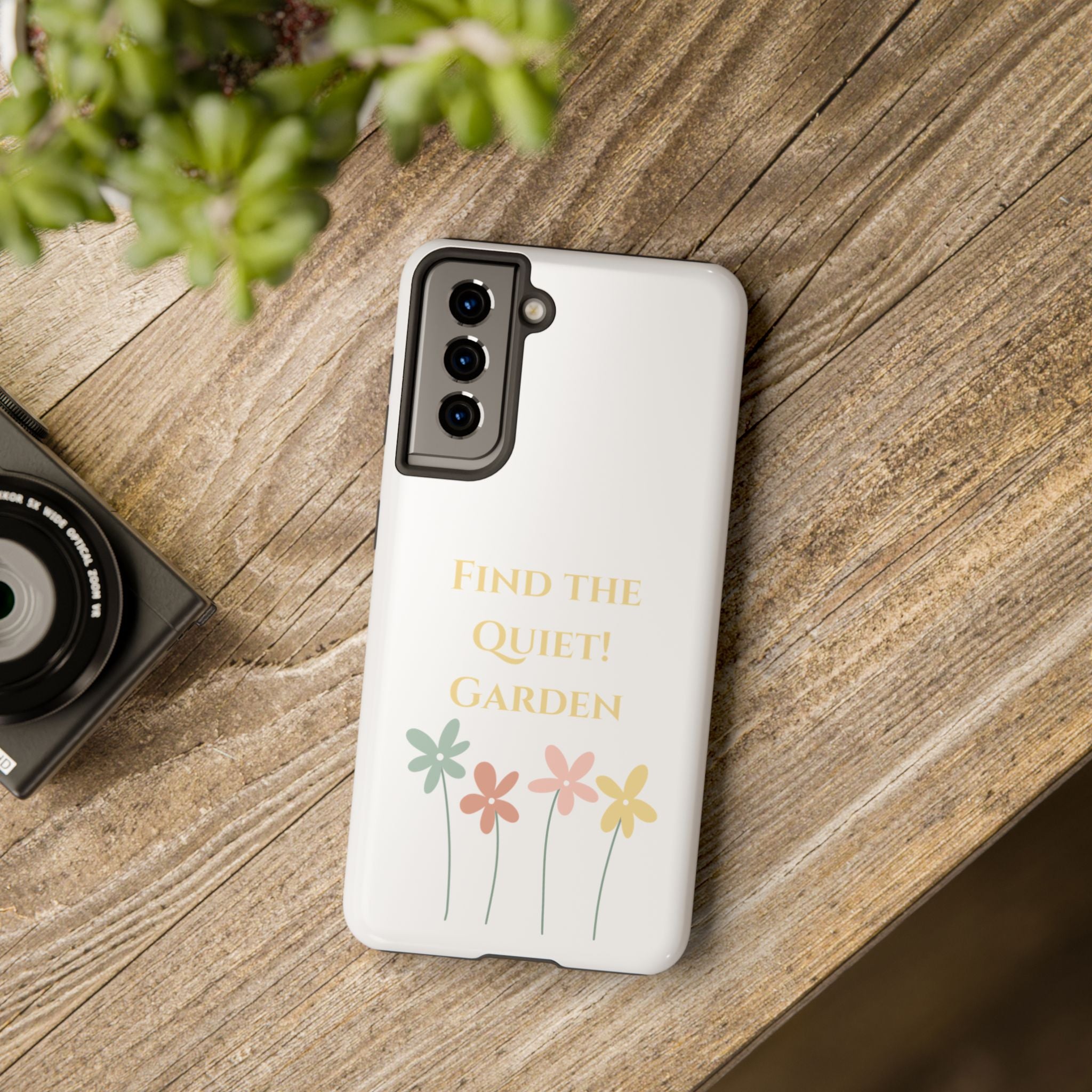 Phone case with floral design and text on a wooden surface with a camera and plant.