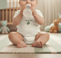 Baby in a white onesie with green text and leaf design, sitting on a mat and drinking from a bottle.