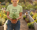 Woman holding a potted plant in a garden wearing a 'Mommy Gardener' t-shirt.