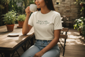 Woman sitting outdoors at a wooden table, holding a mug and wearing a white t-shirt with text.