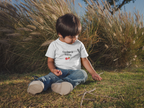Child sitting in grass wearing a white t-shirt with text and graphics