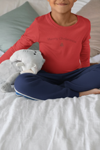 Child wearing a red 'Merry Christmas' shirt sitting on a bed with a gray pillow and white blanket.