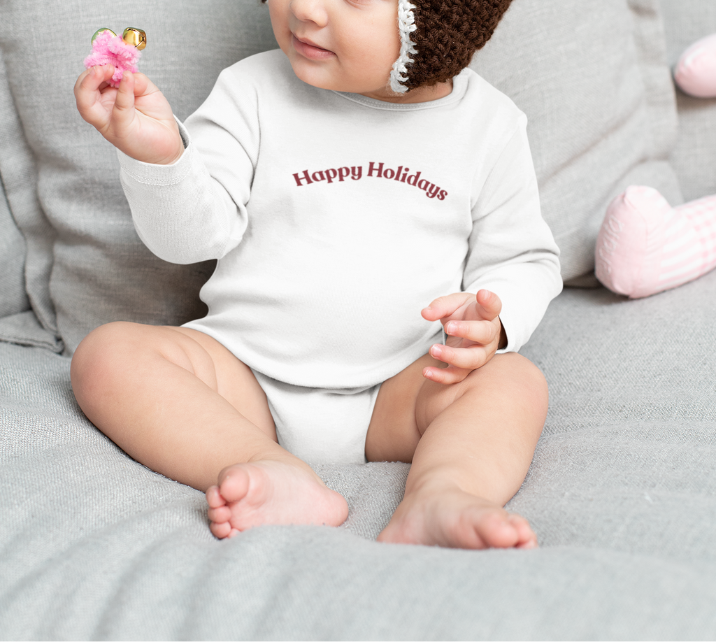 Baby wearing a brown crochet hat with a white flower and a 'Happy Holidays' onesie, sitting on a gray couch.