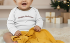 Baby wearing a 'Happy Holidays' onesie sitting on a yellow blanket with Christmas decorations in the background.