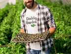 Man holding a tray of plants in a greenhouse with 'Daddy Garden' shirt