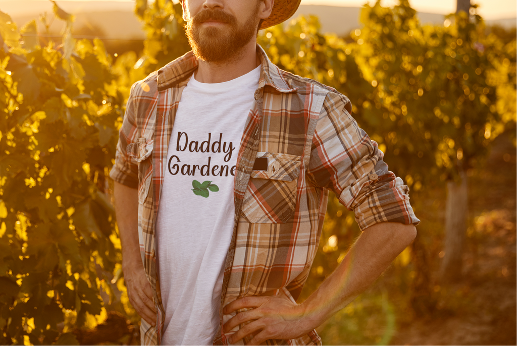 Man wearing a hat and plaid shirt with 'Daddy Garden' t-shirt in a vineyard at sunset.