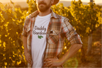 Man wearing a hat and plaid shirt with 'Daddy Garden' t-shirt in a vineyard at sunset.