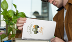 Notebook with a garden-themed design on a wooden surface with a cup of coffee and plant.