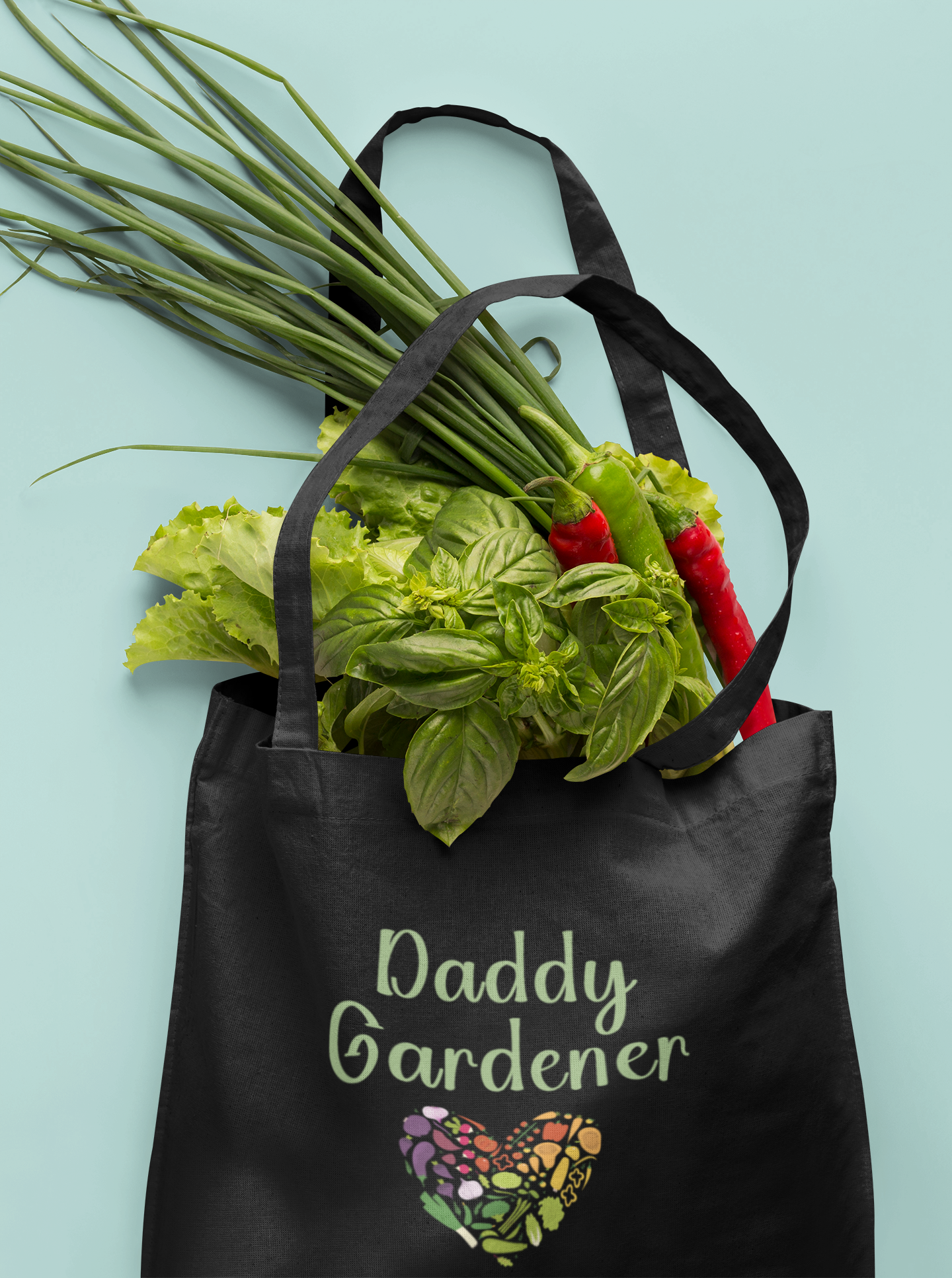 Black tote bag with 'Daddy Gardener' text and heart design, filled with fresh vegetables on a light blue background.