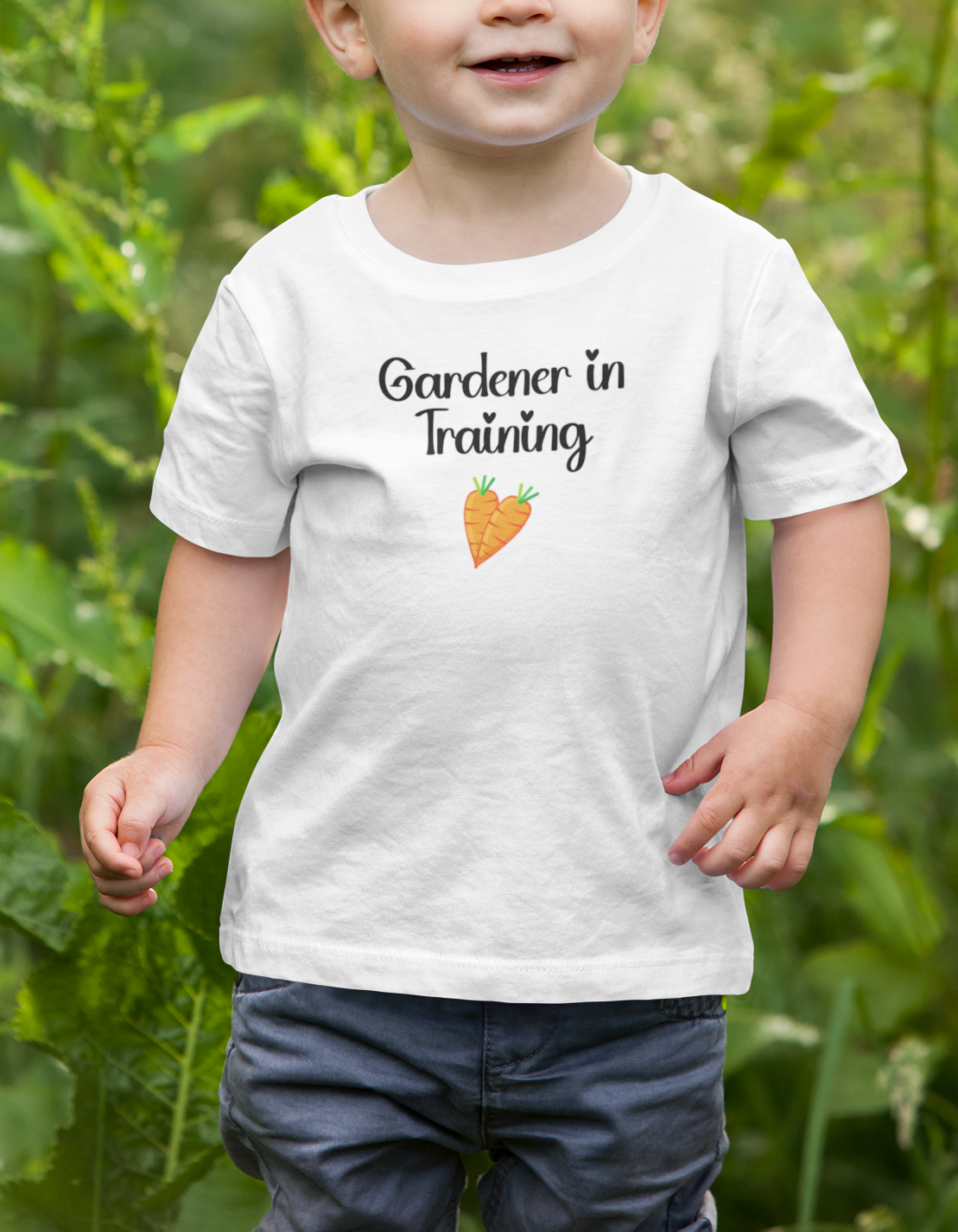 Child wearing a white t-shirt with 'Gardener in Training' and a carrot graphic, standing in a garden.