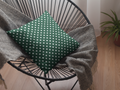 Green pillow with white polka dots on a black wire chair with a gray blanket and plant in the background.