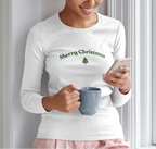 Woman wearing a 'Merry Christmas' shirt, holding a mug and phone, against a light-colored wall.