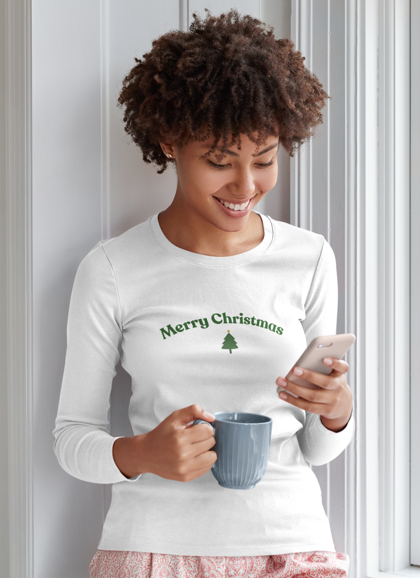 Woman wearing a 'Merry Christmas' shirt, holding a mug and phone, against a light-colored wall.