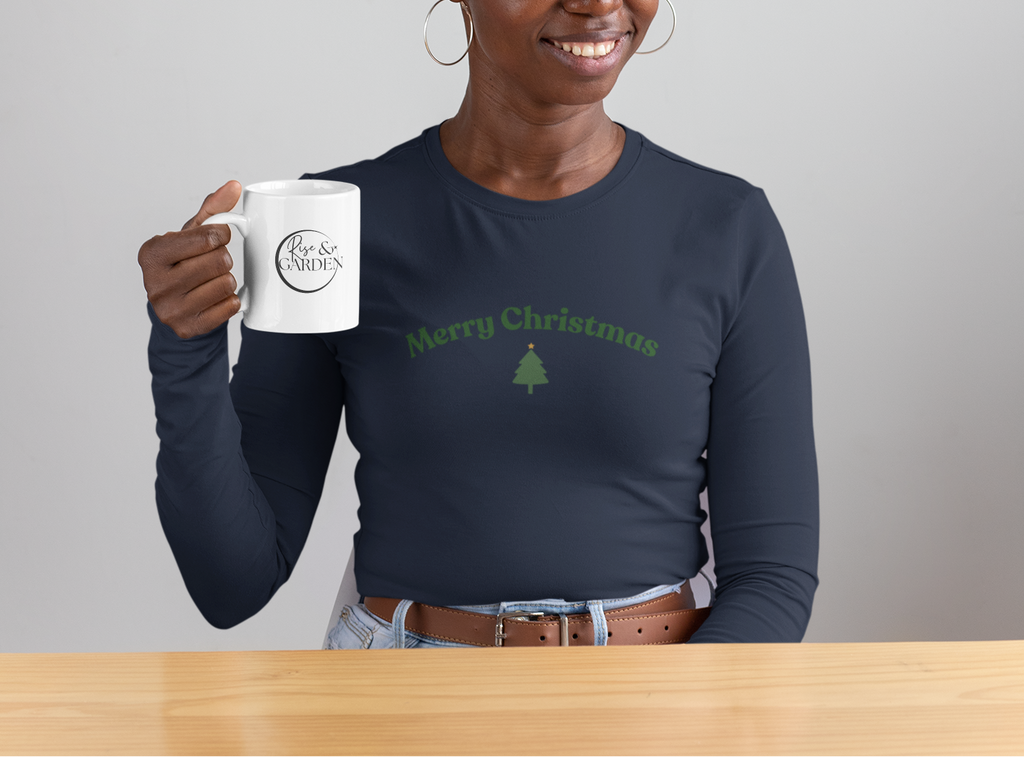 Person holding a mug with a Christmas-themed design, wearing a navy blue shirt with a green tree and text, in front of a festive wreath.