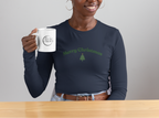 Person holding a mug with a Christmas-themed design, wearing a navy blue shirt with a green tree and text, in front of a festive wreath.