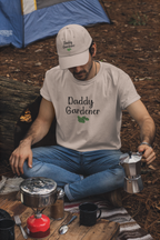 Man in 'Daddy Gardener' t-shirt sitting outdoors with camping gear and coffee equipment.