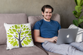 Man using a laptop on a couch with a decorative pillow featuring a tree design.