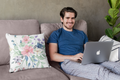 Man using a laptop on a couch with decorative pillows and a plant in the background