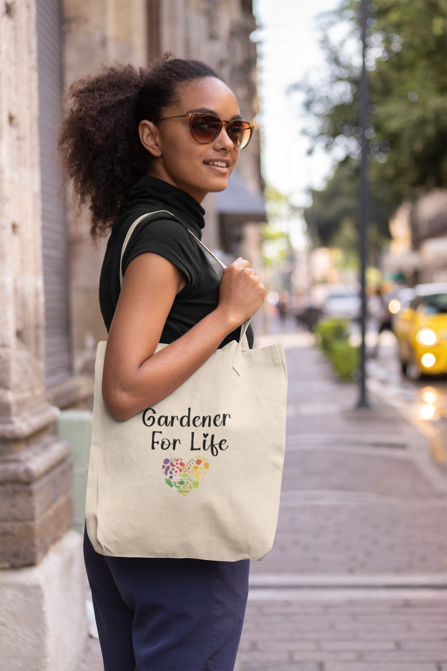 Woman holding a tote bag with 'Gardener For Life' text on a city street.