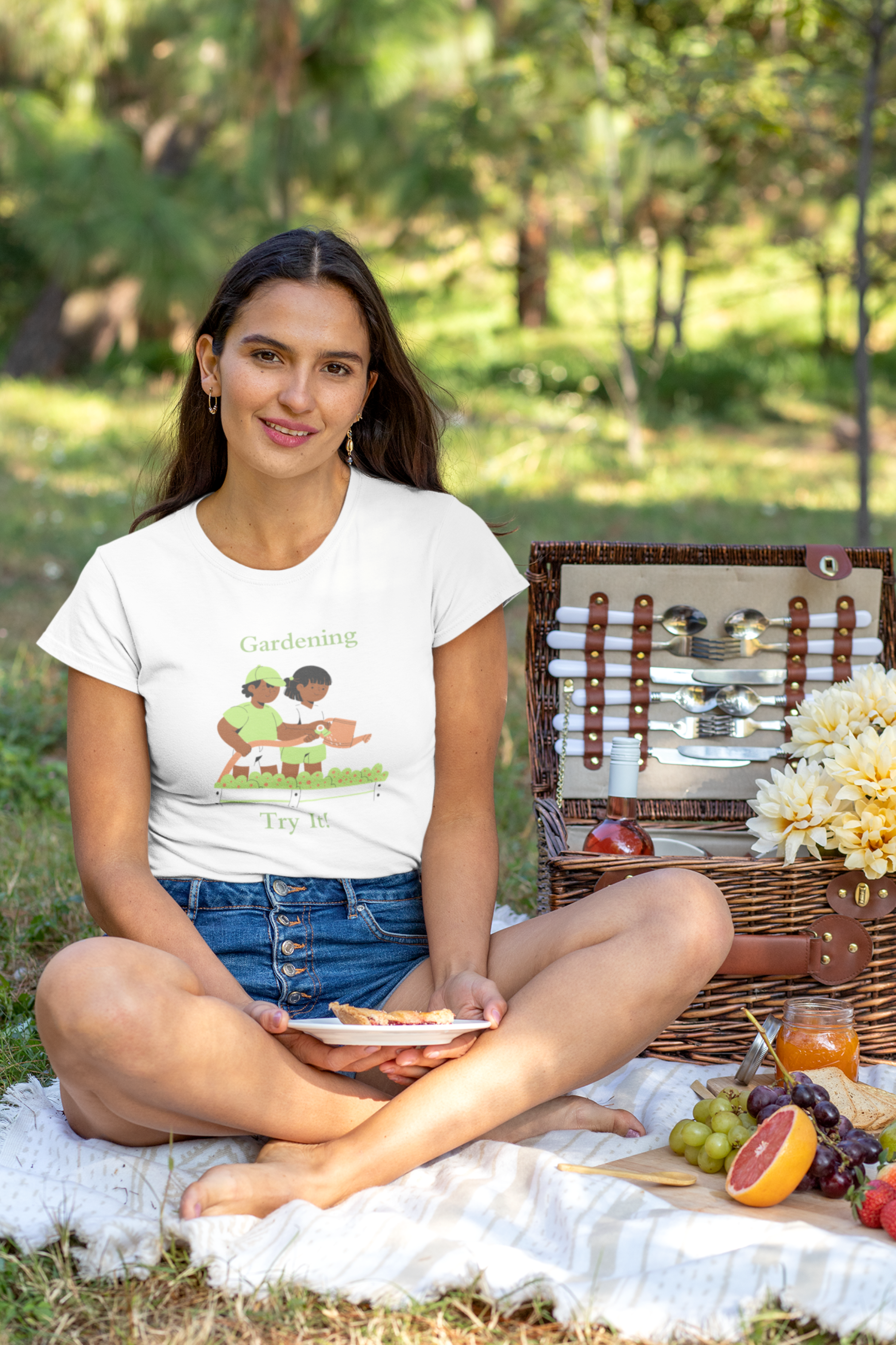 Woman sitting on a picnic blanket with a basket and food, wearing a white t-shirt with a graphic design.