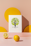 Framed artwork with a tree design and text, placed on a pink surface with lemons and yellow circular shapes.