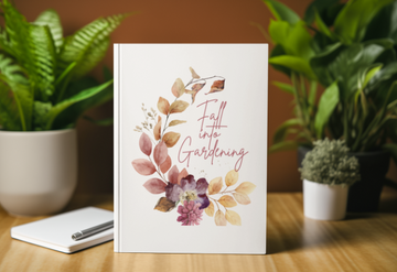 Greeting card with floral design and text 'Fall into Gardening' on a wooden table with plants in the background.