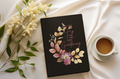 Black notebook with floral design and text on a white surface with flowers and a cup of coffee.