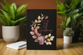 Notebook with floral design and text 'Fall into Gardening' on a wooden surface with plants in the background