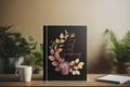 Black notebook with floral design and text on a wooden table with plants in the background